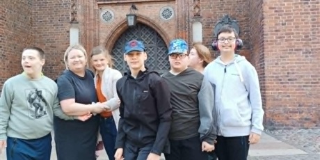 Powiększ grafikę: Grupa chłopców, dziewczynka i dwie kobiety stoją na ulicy. W tle zabytkowy kościół i fragment flagi Polski.