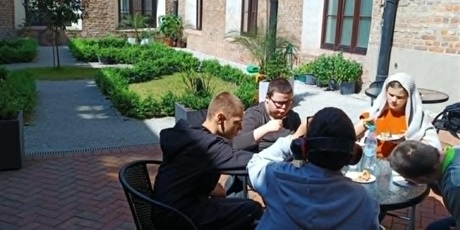 Powiększ grafikę: Grupa dzieci siedzi przy stoliku i je ciasto. W tle zabytkowy dziedziniec