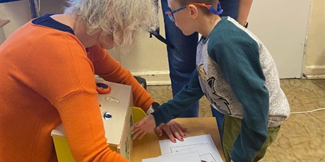 Powiększ grafikę: Na pierwszym planie dziecko wkłada rękę do skrzynki z ukrytymi darami jesieni. Kobieta trzyma skrzynkę i rozkłada symbole PCS do wskazania co dziecko dotyka w skrzynce bez kontroli wzroku.