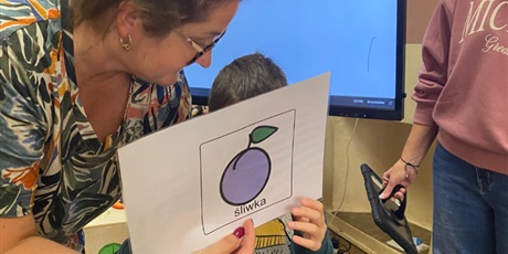 Powiększ grafikę: W tle ekran multimedialny oraz kobieta, która trzyma komunikator Mówik. Na pierwszym planie dziecko pokazuje wszystkim symbol PCS ćliwka. Druga kobieta pomaga dziecku przytrzymać symbol.