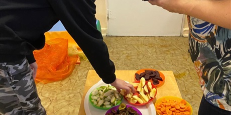 Powiększ grafikę: W tle szyfony i chusty w jesiennych kolorach. Na pierwszym planie taca do degustacji. Talerzyki z pokrojonymi jabłkami, śliwkami, gruszkami, marchewkami, gotowanymi buraczkami. Chłopiec sięga po jabłk