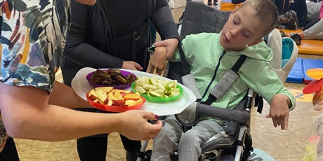 Powiększ grafikę: W tle siedzą dzieci na materacach i klockach sensorycznych. Chłopiec na wózku inwalidzkim próbuje wybrać z talerzyków jabłek, gruszek i śliwek co chce zjeść. Jedna kobieta trzyma tacę. Druga pomaga uc