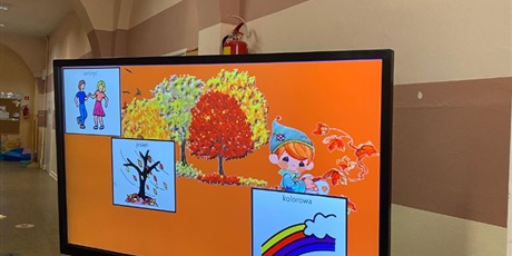 Powiększ grafikę: Tablica multimedialna z prezentacją AAC do piosenki "Tańcowała jesień". Pomarańczowe tło prezentacji, kolorowe dzrewa w tle, symbole obrazkowew tańczyć, jesień, kolorowa.