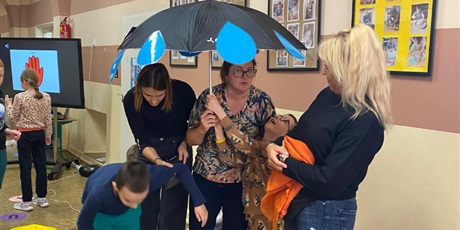 Powiększ grafikę: W tle tablica multimedialna i sylwetki dzieci. Na pierwszym planie dwie kobiety i dwójka dzieci chowają się pod parasol z przyczepionymi dużymi kroplami deszczu. Przygląda się im kobieta.