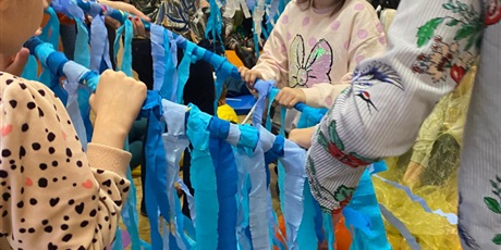 Powiększ grafikę: W tle widać dzieci tańczące w grupkach z kółkami hulahop. Na pierwszym planie dwie kobiety i dwójka dzieci trzymają hulahop z niebieskimi paskami z bibuły i tańczą.