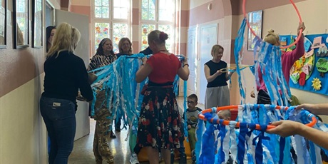 Powiększ grafikę: Zdjęcie zbiorowe. Dzieci w grupkach, wspierane przez panie tańczą ze swoimi kółkami hulahop i machają przyczepionymi paskami bibuły.