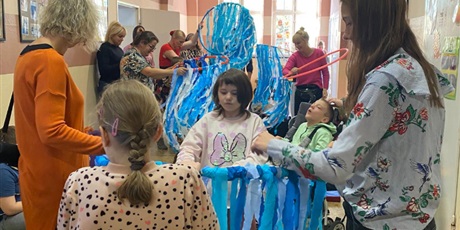 Powiększ grafikę: Zdjęcie zbiorowe. Dzieci w grupkach, wspierane przez panie tańczą ze swoimi kółkami hulahop i machają przyczepionymi paskami bibuły.W tle przygląda im się chłopiec na wózku.