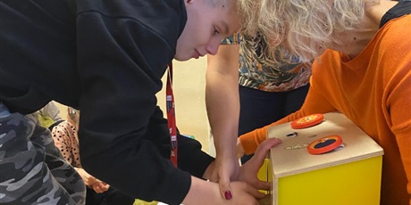 Powiększ grafikę: Na pierwszym planie chłopiec próbuje włożyć rękę do skrzyneczki skrytki z darami jesieni. Jedna pani trzyma skrzynkę, druga pomaga włożyć mu rękę w przeznaczony do tego otwór. 