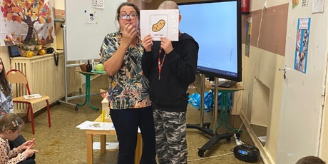 Powiększ grafikę: W tle tablica multimedialna, tablica z symbolami obrazkowymi, stoliki z warzywami. Na pierwszym planie kobieta obok chłopca, który pokazuje wszystkim symbol PCS ziemniak.