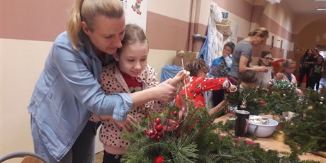 Powiększ grafikę: Kobieta i dziewczynka przygotowują stroik świąteczny. W tle grupa ludzi pracujących przy stole i przygotowujących świąteczne stroiki.       