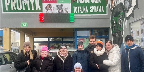 Powiększ grafikę: 4.	Grupa ludzi pozuje do zdjęcia. W tle widac budynek z nazwą schroniska