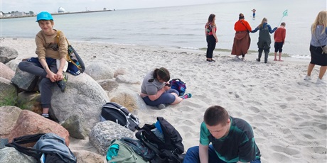 Powiększ grafikę: Uczniowie siedzą na plaży i odpoczywają, na drugim planie z uczniami przy brzegu morza stoi wychowawczyni z uczniami.