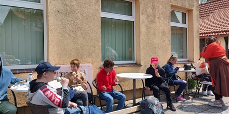 Powiększ grafikę: Dzieci i chłopcy siedzą przy stole w ogródku kawiarnianym i jedzą lody. W tle budynki