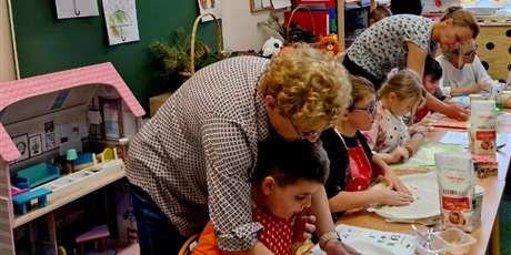 Powiększ grafikę: 9.	Dzieci siedzą przy stolikach, a  za nimi stoją panie i  pomagają im przy wyrabianiu ciasta.