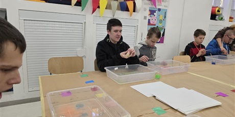 Powiększ grafikę: Chłopcy siedzą przy stole, na którym są pojemniki z cieczą i patyczkami do szaszłyków nakrapiają  farbę na powierzchnię cieczy.