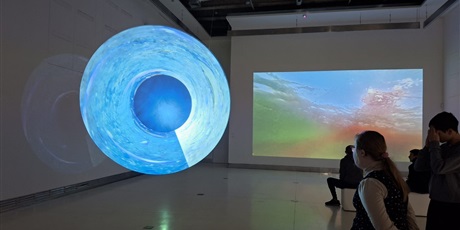 Powiększ grafikę: W zaciemnionej sali uczniowie obserwują świetlny duży okrąg w kolorze niebieskim. W tle ekran z kolorową abstrakcją.