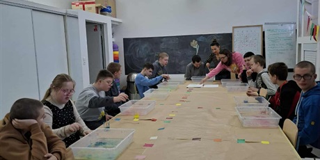 Powiększ grafikę: Chłopcy siedzą przy stole, na którym są pojemniki z cieczą i patyczkami do szaszłyków nakrapiają  farbę na powierzchnię cieczy..w Tle zieloan tablica i szafy.