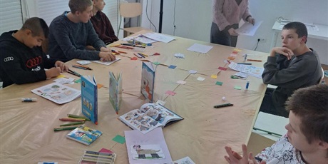 Powiększ grafikę: Pięciu uczniów siedzi za stołem konferencyjnym i ogląda prelekcję na ekranie projekcyjnym. Pod ekranem prelegentka pokazuje kartki papieru.