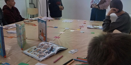 Powiększ grafikę: Pięciu uczniów siedzi za stołem konferencyjnym i ogląda prelekcję na ekranie projekcyjnym. Pod ekranem prelegentka pokazuje kartki papieru. Obok niej stoi uczeń