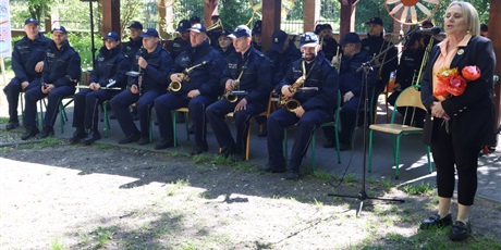 Powiększ grafikę: Orkiestra w mundurach, z instrumentami siedzi na krzesłach pod wiatą, po prawej stronie nauczycielka (dyrektorka) wygłasza przemówienie do mikrofonu, w tle ogrodzenie Ośrodka