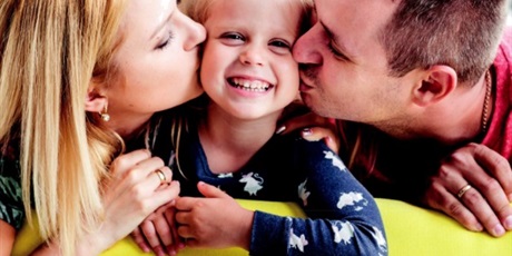 Powiększ grafikę: Dwoje rodziców całuje w policzki swoje uśmiechnięte dziecko